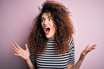 Young beautiful woman with curly hair and piercing wearing casual striped t-shirt crazy and mad shouting and yelling with aggressive expression and arms raised. Frustration concept.