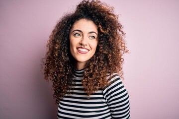 Young beautiful woman with curly hair and piercing wearing casual striped t-shirt looking away to side with smile on face, natural expression. Laughing confident.