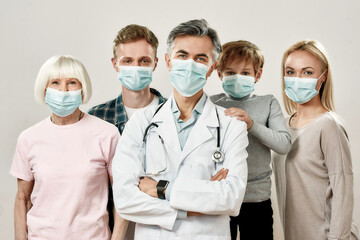 Family Physician. Portrait of mature male doctor and family of four wearing medical protective masks, looking at camera while standing against grey background
