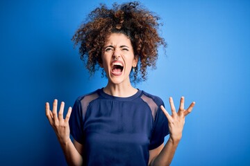 Young beautiful woman with curly hair and piercing wearing casual blue t-shirt crazy and mad shouting and yelling with aggressive expression and arms raised. Frustration concept.