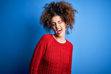 Young beautiful woman with curly hair and piercing wearing casual red sweater winking looking at...