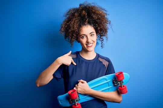 Young sporty woman with curly hair and piercing holding skate over blue background with surprise face pointing finger to himself
