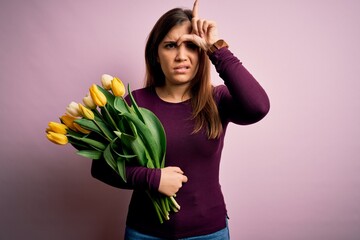 Young blonde woman holding romantic bouquet of yellow tulips flowers over pink background making fun of people with fingers on forehead doing loser gesture mocking and insulting.