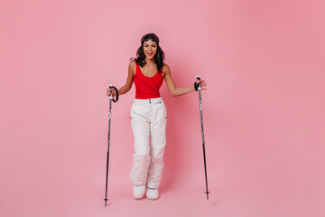 Smiling adorable woman posing with ski poles. Full length view of girl in white pants standing on pink background.
