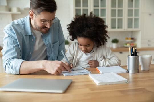 Caring Caucasian stepfather help with homework adorable mixed-race daughter. Home tutor teaches school subject to little learner, sitting together in domestic kitchen. Education, homeschooling concept