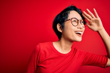 Young beautiful asian girl wearing casual t-shirt and glasses over isolated red background very...