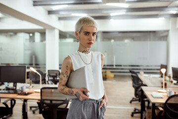 I am ready to work. Young, stylish and attractive tattooed business woman holding her laptop and looking at camera while standing in the modern coworking space