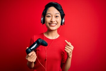 Young beautiful asian gamer girl playing video game using joystick and headphones very happy pointing with hand and finger © Krakenimages.com