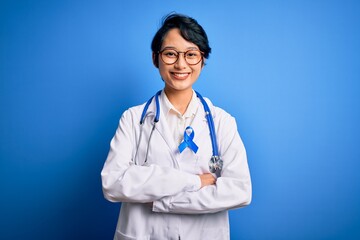 Young beautiful asian doctor girl wearing stethoscope and coat with blue cancer ribbon happy face smiling with crossed arms looking at the camera. Positive person.