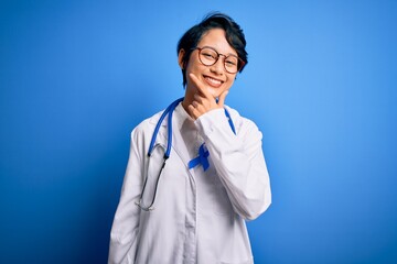Young beautiful asian doctor girl wearing stethoscope and coat with blue cancer ribbon looking confident at the camera smiling with crossed arms and hand raised on chin. Thinking positive.