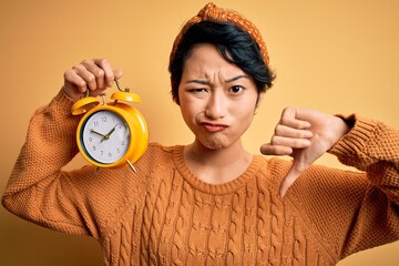 Young beautiful chinese woman holding vintage alarm clock over isolated yellow background with...