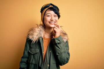 Beautiful asian skier girl wearing snow sportswear using ski goggles over yellow background looking confident at the camera with smile with crossed arms and hand raised on chin. Thinking positive.
