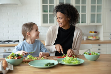 African mother teach Caucasian small cute daughter preparing healthy vegetarian food cutting vegetables, mum shares salad recipe, family chatting enjoy time and hobby in kitchen, cook together concept