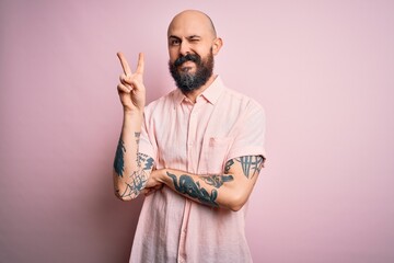 Handsome bald man with beard and tattoo wearing casual shirt over isolated pink background smiling with happy face winking at the camera doing victory sign. Number two.