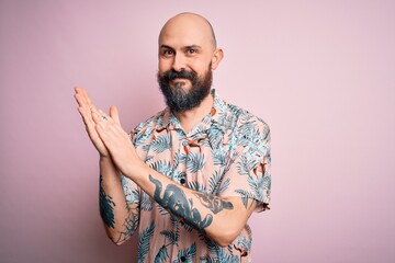 Handsome bald man with beard and tattoo wearing casual floral shirt over pink background clapping and applauding happy and joyful, smiling proud hands together