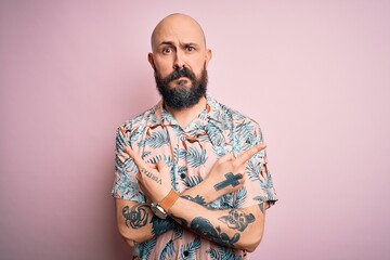 Handsome bald man with beard and tattoo wearing casual floral shirt over pink background Pointing to both sides with fingers, different direction disagree