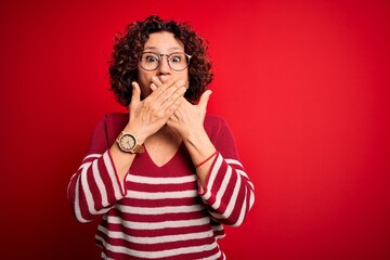 Middle age beautiful curly hair woman wearing casual striped sweater over red background shocked covering mouth with hands for mistake. Secret concept.