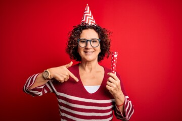 Obraz premium Middle age curly hair woman wearing birthday funny hat holding party trumpet on celebration with surprise face pointing finger to himself