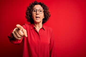 Middle age beautiful curly hair woman wearing casual shirt and glasses over red background pointing...