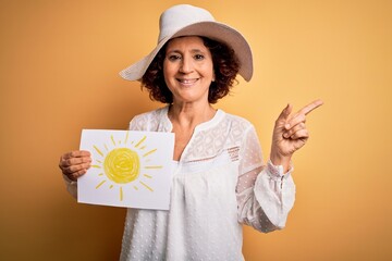 Middle age curly woman on vacation holding bunner with sun image over yellow background very happy pointing with hand and finger to the side