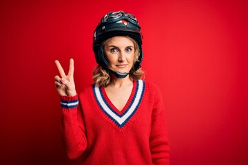 Middle age beautiful blonde motorcyclist woman wearing moto helmet over red background smiling with happy face winking at the camera doing victory sign. Number two.