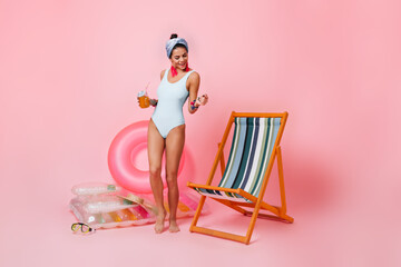 Joyful young woman in white swimsuit dancing near chaise longue. Studio shot of magnificent girl with cocktail standing on pink background.