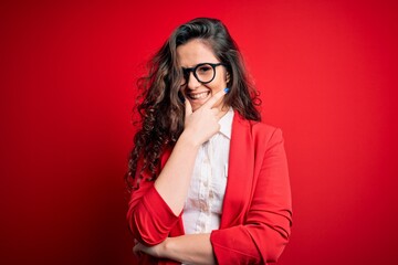 Young beautiful woman with curly hair wearing jacket and glasses over red background looking confident at the camera smiling with crossed arms and hand raised on chin. Thinking positive.