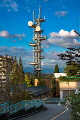 Obraz premium A telecommunications tower located in uptown of New Westminster among buildings and trees against a mountain range and a beautiful cloudy sky. New Westminster City, British Columbia, Canada