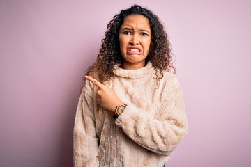 Young beautiful woman with curly hair wearing casual sweater standing over pink background Pointing aside worried and nervous with forefinger, concerned and surprised expression