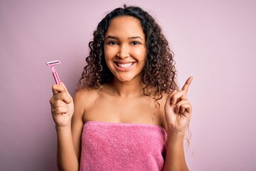 Young beautiful woman with curly hair wearing shower towel holding depilation razor surprised with...
