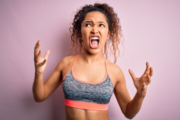 Beautiful sportswoman with curly hair doing sport wearing sportswear over pink background crazy and mad shouting and yelling with aggressive expression and arms raised. Frustration concept.