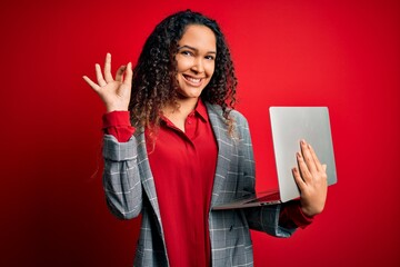 Young beautiful business woman with curly hair working using laptop over red background doing ok...