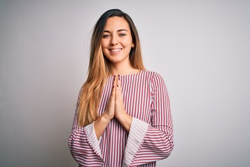 Young beautiful blonde woman with blue eyes wearing stiped t-shirt over white background praying with hands together asking for forgiveness smiling confident.