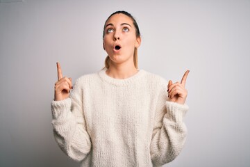 Young beautiful blonde woman with blue eyes wearing casual sweater over white background amazed and surprised looking up and pointing with fingers and raised arms.