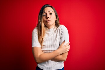 Beautiful blonde woman with blue eyes wearing casual white t-shirt over red background shaking and freezing for winter cold with sad and shock expression on face