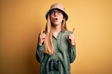 Beautiful blonde explorer woman with blue eyes wearing hat and glasses using binoculars amazed and surprised looking up and pointing with fingers and raised arms.