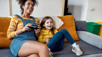 The best care taker. African american woman, baby sitter and little girl having fun together, playing video games, sitting on the couch. Children education, leisure activities, babysitting concept