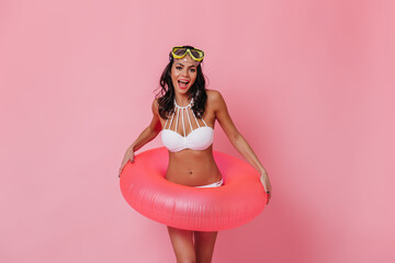 Joyful girl in bikini standing on pink background. Smiling brunette woman posing with swimming circle.