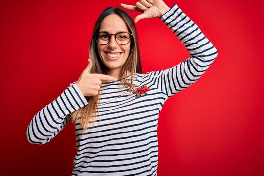 Young beautiful blonde woman with blue eyes wearing glasses standing over red background smiling making frame with hands and fingers with happy face. Creativity and photography concept.