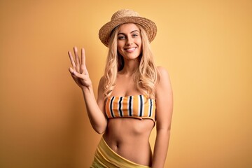 Young beautiful blonde woman on vacation wearing bikini and hat over yellow background showing and...