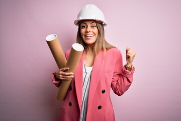 Young beautiful brunette architect woman wearing safety helmet holding blueprints screaming proud...