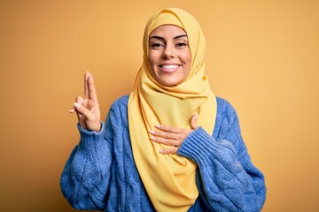 Young beautiful brunette muslim woman wearing arab hijab over isolated yellow background smiling...