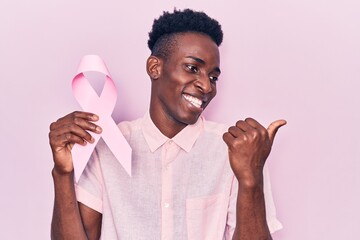 Young african american man holding pink cancer ribbon pointing thumb up to the side smiling happy...