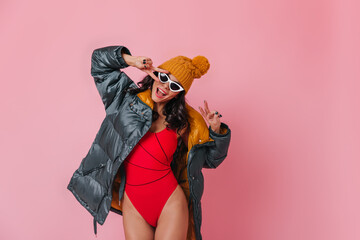 Appealing girl in swimsuit and hat posing with peace signs. Studio shot of laughing caucasian woman...