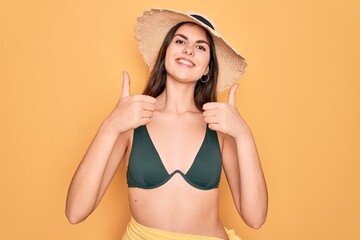 Young beautiful girl wearing swimwear bikini and summer sun hat over yellow background approving doing positive gesture with hand, thumbs up smiling and happy for success. Winner gesture.