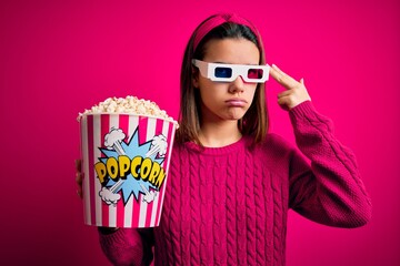 Obraz premium Young beautiful girl watching movie using 3d glasses eating box with popcorns Shooting and killing oneself pointing hand and fingers to head like gun, suicide gesture.