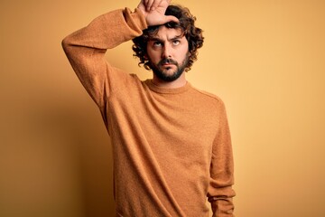 Young handsome man with beard wearing casual sweater standing over yellow background making fun of people with fingers on forehead doing loser gesture mocking and insulting.