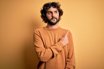 Young handsome man with beard wearing casual sweater standing over yellow background Pointing with hand finger to the side showing advertisement, serious and calm face