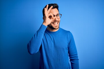 Young handsome man with beard wearing casual sweater and glasses over blue background doing ok gesture with hand smiling, eye looking through fingers with happy face.