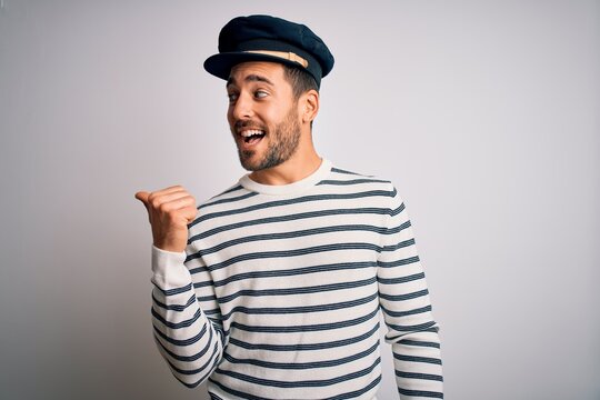 Young handsome sailor man with beard wearing navy striped uniform and captain hat smiling with happy face looking and pointing to the side with thumb up.